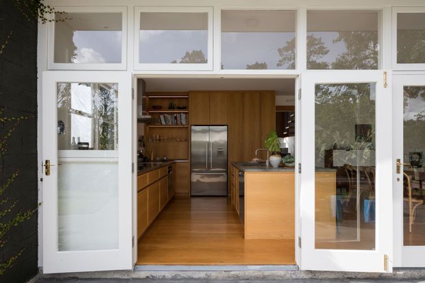 View into a warm timber-finished kitchen through a white-framed glazed folding or bi-fold door opening. The kitchen features wood cabinetry, a central island, stainless-steel appliances, open shelving, and timber flooring. Reflections of surrounding trees are visible in the extensive glazing, while dark exterior cladding and climbing greenery frame the entrance.