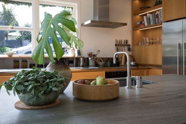 Warm, light-filled contemporary kitchen with a large central island in a dark grey wood-look finish, timber cabinetry, open shelving, stainless-steel appliances, and a sculptural stainless-steel tap and sink. Large windows bring in natural light, while potted greenery and a timber fruit bowl add an organic accent.