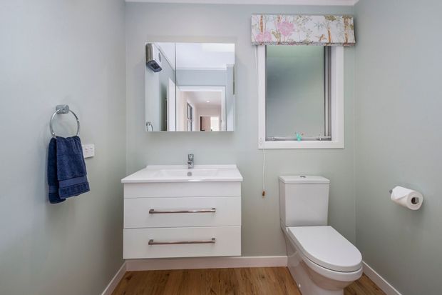 A compact, clean bathroom with pale grey-green walls, white wall-mounted vanity and basin, chrome tapware, toilet, framed frosted window, floral fabric pelmet, towel ring with navy towels, and warm timber flooring.