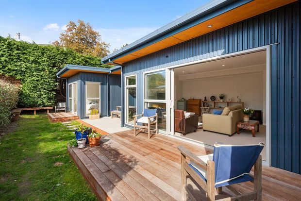 Exterior view of a contemporary single-storey home opening onto a timber deck and landscaped garden. The building features dark blue-grey vertical metal cladding, a low-pitched roof with broad timber-lined eaves, large glazed sliding doors, and a bright living room visible through the opening. Outdoor seating and potted plants occupy the deck.