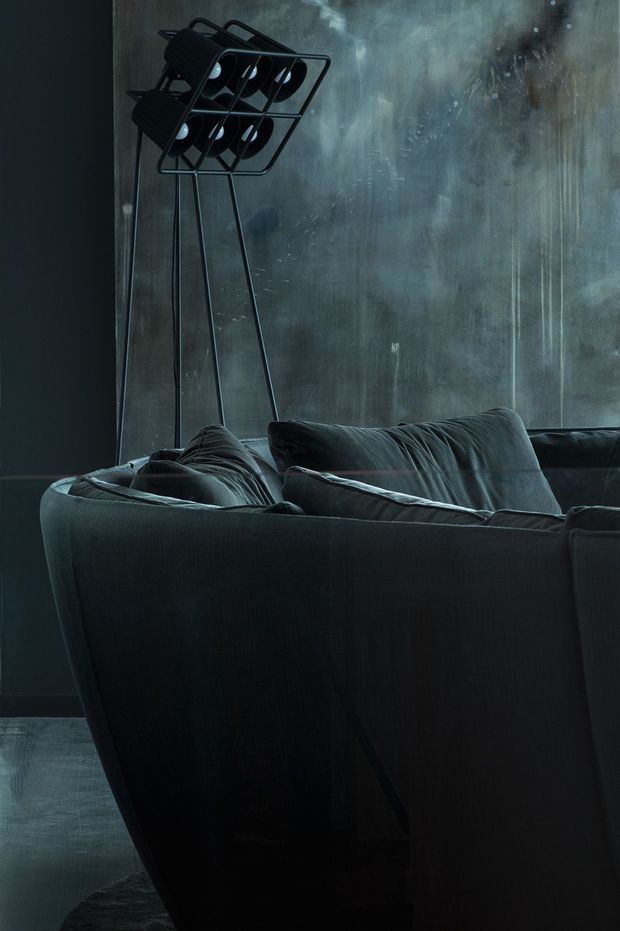 Moody interior view of a dark upholstered sofa with soft cushions, set against a distressed blue-grey textured wall. A tall black industrial-style multi-lamp floor light stands behind the sofa, and a dark floor covering is visible below.