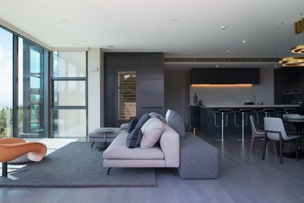 Contemporary open-plan living and kitchen area with floor-to-ceiling glazing along one side, a large pale sectional sofa, dark built-in cabinetry, a compact kitchen island with bar stools, and a dining area. The space has a restrained grey, charcoal, and pale neutral palette with layered pendant lighting and a bright outlook.