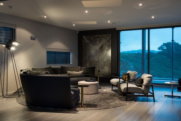A sophisticated contemporary living room with dark upholstered sofas and armchairs arranged around low round coffee tables on a large rug. The room has grey walls, warm timber flooring, recessed ceiling lighting, a dramatic dark feature wall with artwork, and expansive floor-to-ceiling glazing overlooking a wooded mountainous landscape.