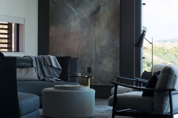 Moody contemporary living room with a deep grey sectional sofa, a rounded upholstered ottoman, and a dark-framed lounge chair. A large distressed dark metallic or concrete-look feature wall dominates the background, while floor-to-ceiling glazing on the right opens to a natural landscape. The palette is charcoal, grey, and muted neutral tones with strong contrast and soft daylight.