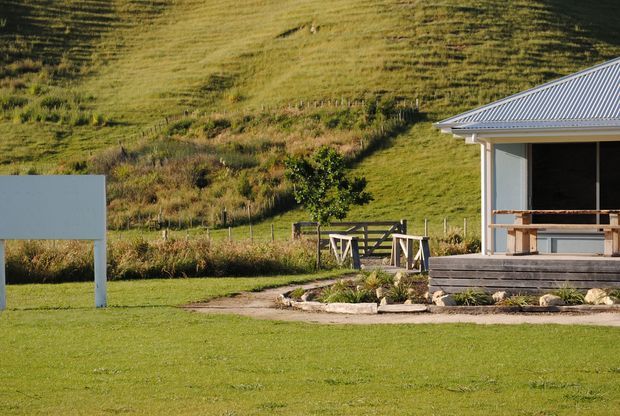 A rural single-storey building viewed from outside, with a simple light-coloured facade, corrugated metal pitched roof, and covered timber-deck porch. The building sits beside a broad mown lawn and landscaped garden, with a fenced grassy hillside and farm-style gate in the background.