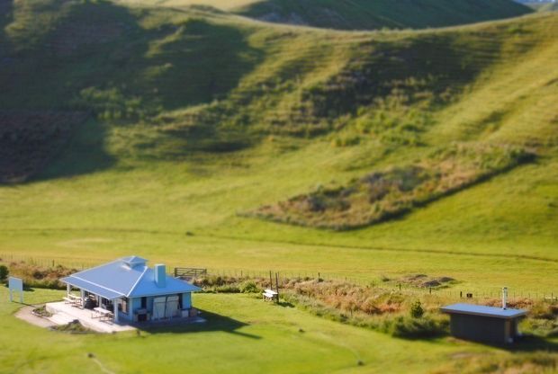 A small modern rural dwelling with a low-pitched roof sits in an expansive green grassy landscape, accompanied by a detached outbuilding. Rolling hills and open pasture dominate the background.