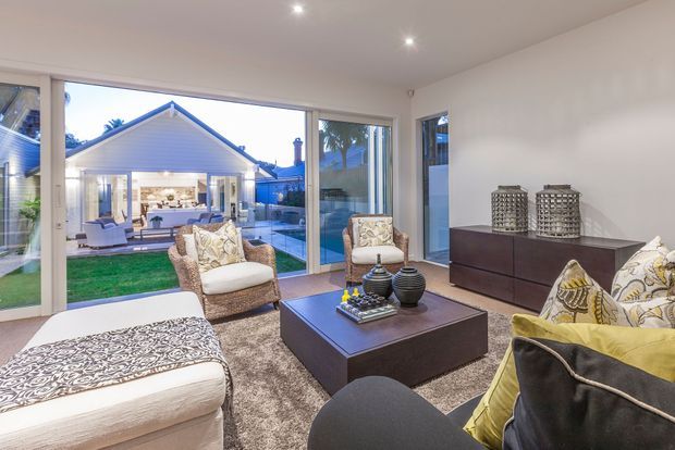 A bright contemporary living room with full-height glazing overlooking a landscaped backyard and a separate gabled pavilion. The room features a large sectional sofa, upholstered armchair, ottoman, dark wood coffee table and sideboard, patterned cushions, and recessed ceiling lighting.
