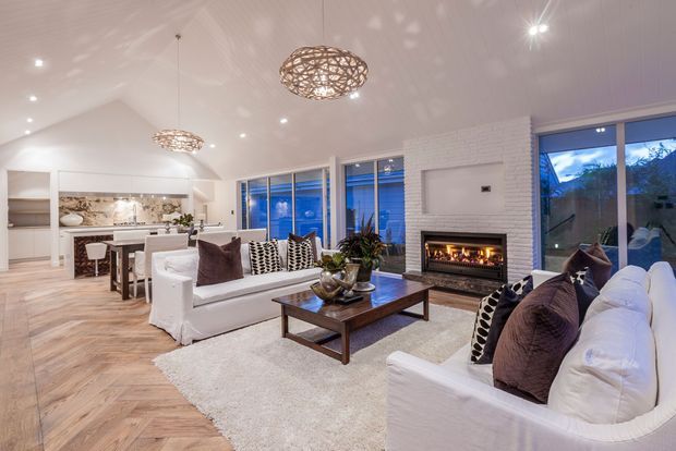 Bright open-plan living and dining area with a white brick fireplace, wide glazing, white slipcovered sofas, patterned cushions, a timber coffee table, pale timber flooring, and woven pendant lights. The adjoining kitchen and dining space are visible in the background, with an outdoor landscape seen through the windows.