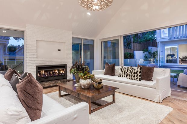 Bright contemporary living room with white sofas, a dark timber coffee table, a white-painted brick fireplace with inset gas fire, pale timber flooring, a textured neutral rug, and large windows and glazed doors opening toward an outdoor area. A woven pendant light and patterned cushions add warm decorative accents.