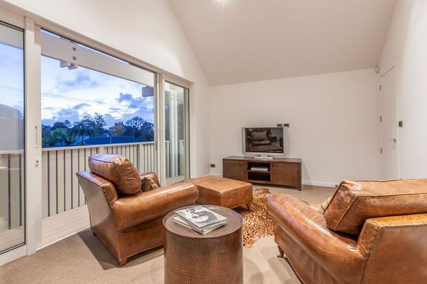 Bright living room with warm tan leather armchairs and sofa arranged around a round ottoman, a patterned area rug, and a compact timber media console with television. Large sliding glass doors open to a balcony, bringing in views and natural light. The room has plain off-white walls, a sloped ceiling, and beige carpet.