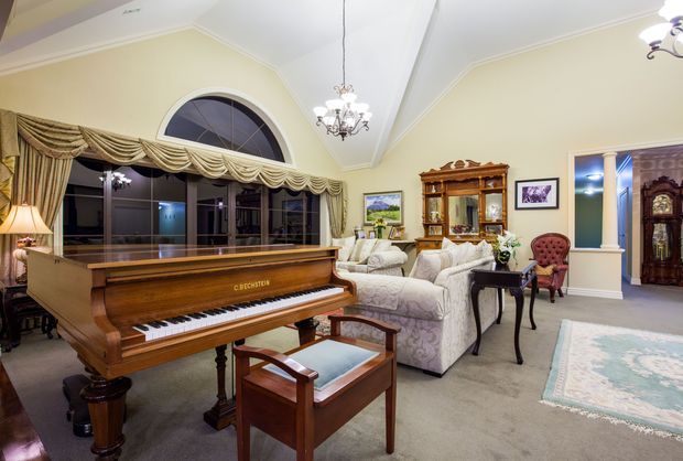 A traditional, warmly lit living room centred around a polished grand piano, with upholstered seating, a display cabinet, ornate chandelier, arched window, draped curtains, framed artwork, and carpeted flooring. The room opens toward a hallway or adjoining space on the right.