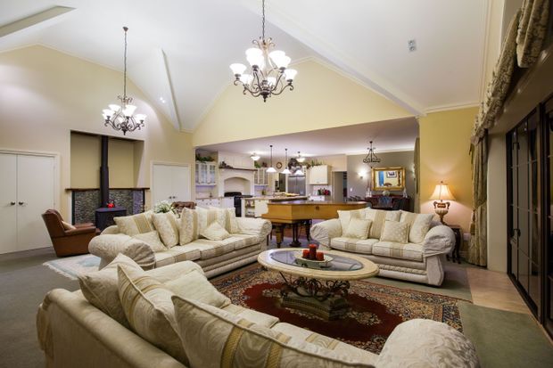 A spacious, traditionally styled open-plan living room with multiple cream upholstered sofas arranged around an ornate oval coffee table and patterned Persian-style rug. The room features pale yellow walls, white decorative ceiling details, several ornate chandeliers, built-in cabinetry, a fireplace area, and an open view into a kitchen and bar space. Tall dark-framed glazing or doors line the right side, with drapery and a table lamp adding warmth.