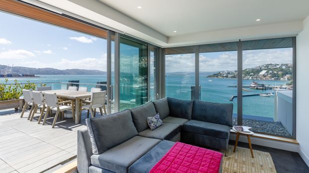Bright contemporary living room with a large grey sectional sofa and ottoman beside full-height corner glazing, opening onto a timber deck with outdoor dining. The windows provide expansive waterfront views over a blue bay and surrounding hills.