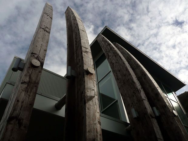 Low-angle exterior view of a contemporary building with dramatic tall, rough-textured timber structural posts or fins rising in front of large angled glazing. The facade combines dark timber, grey-green metal cladding, exposed cylindrical fixtures, and a pronounced projecting roof edge beneath a cloudy sky.