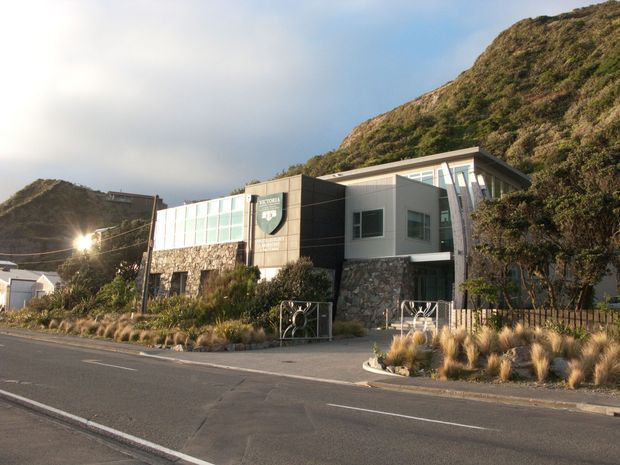 A contemporary multi-level building set against a steep vegetated hillside, viewed from across a paved road. The facade combines dark rectangular cladding, pale rendered walls, extensive glazed panels, and irregular natural stonework. Native-style grasses and shrubs line the frontage, with a low metal fence and pedestrian entrance. The building has a low-pitched roof and a prominent projecting upper volume.