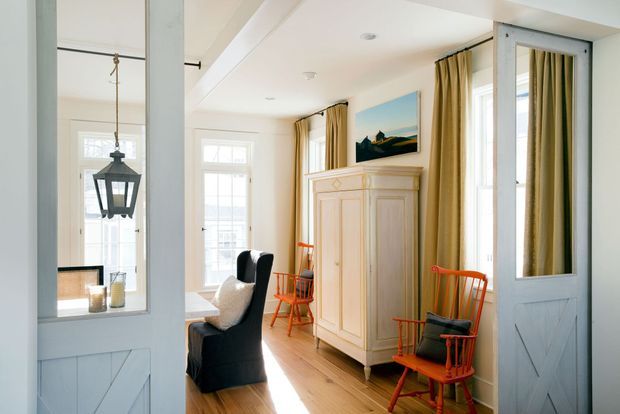 Bright, narrow traditional-style sitting room or hallway with white-painted timber walls and doors, warm wood flooring, tall windows dressed in mustard-gold curtains, a large pale built-in cabinet, black upholstered seating, and distinctive red rocking chairs. A mirrored door reflects a hanging lantern and adjacent seating area.