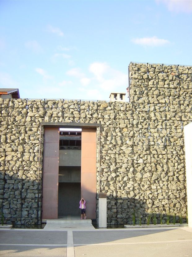 Exterior architectural view of a substantial stone gabion wall facade with a tall recessed entrance framed in warm rust-coloured metal. The layered wall steps upward around the doorway, with glimpses of a modern building behind it. A person at the entrance provides scale. Bright blue sky and paved foreground are visible.