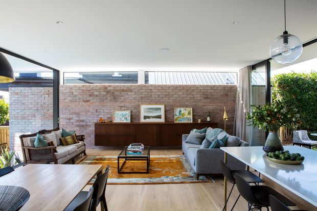 A bright open-plan living and dining area with large black-framed windows, a full-height exposed brick feature wall, and light timber flooring. The room contains a grey sofa, timber-framed lounge chairs, a dark timber sideboard, glass coffee table, patterned area rug, dining table, and a stone-topped kitchen island or breakfast bar. Indoor plants and pendant lighting add warmth, while framed artwork sits on the sideboard.