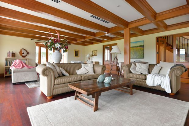 A spacious, traditionally styled living room with warm timber coffered ceiling beams, polished reddish timber flooring, cream upholstered sofas and armchairs, and a large light rug. A substantial dark timber coffee table anchors the seating area, while artwork, a floral arrangement, lamps, and shelving add decorative detail. Glazed doors and an open stair or hallway area are visible in the background.