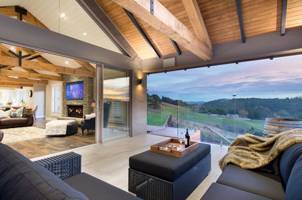 A spacious contemporary living area opening onto a glazed outdoor-facing zone, with a vaulted timber ceiling, exposed heavy timber beams, dark steel supports, and expansive frameless glazing overlooking a green rural landscape. The interior includes dark upholstered sofas, a central ottoman, a stone fireplace visible through the opening, and warm layered lighting.