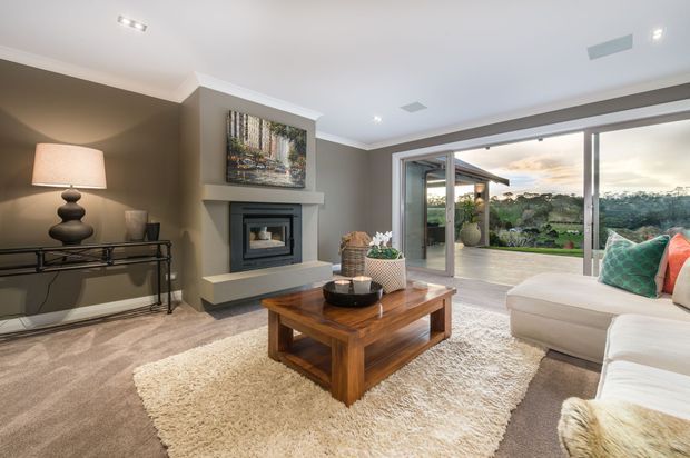 A spacious, softly furnished living room with a neutral taupe and cream palette. The room features a built-in fireplace with a simple mantel and artwork above, a glass-topped console table, a substantial timber coffee table, a large cream shag rug, and a light sectional sofa with colourful cushions. Wide sliding glass doors open to a covered outdoor terrace and landscaped views, creating a bright indoor-outdoor connection.