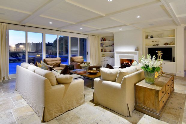 A bright, traditional living room with cream slipcovered sofas and armchairs arranged around a low coffee table and fireplace. Built-in open shelving and cabinetry line the walls, while large glazed doors overlook an outdoor area. The room features a coffered white ceiling, pale stone flooring, and a rustic timber chest used as a side table, creating a warm, refined atmosphere.