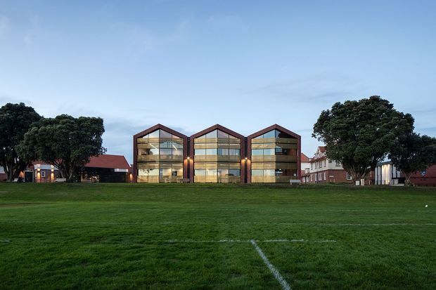 Exterior view of a contemporary multi-storey building comprising three connected gabled volumes with extensive glazed façades and dark structural framing. The building sits beyond a broad grass sports field, with mature trees and older brick buildings surrounding it under an overcast blue evening sky.