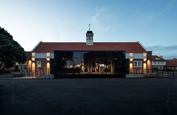 Symmetrical exterior view of a low-rise civic or commercial building with a red tiled pitched roof, central cupola, dark glazed entrance pavilion, and warm brick side wings. The building fronts a broad paved forecourt and is set among other traditional brick buildings at dusk.