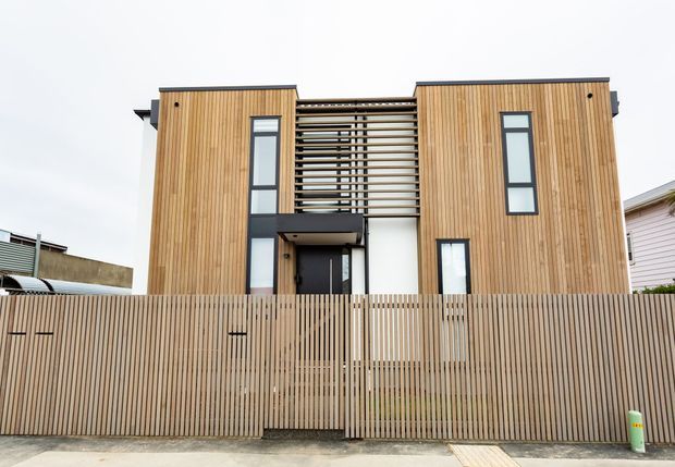Front exterior view of a contemporary multi-storey residence with a symmetrical form, warm vertical timber cladding, dark-framed windows, and a central entrance screened by horizontal timber slats. A vertical timber privacy fence encloses the front boundary.