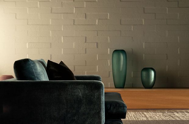 A moody, close-up interior view of a living room. The foreground is dominated by the back and arm of a dark upholstered sofa with cushions, facing a low warm timber media or console cabinet. Two green sculptural ceramic vases sit on the cabinet against a painted white exposed-brick wall. A neutral textured rug is visible at floor level, and directional lighting creates a calm, refined atmosphere.