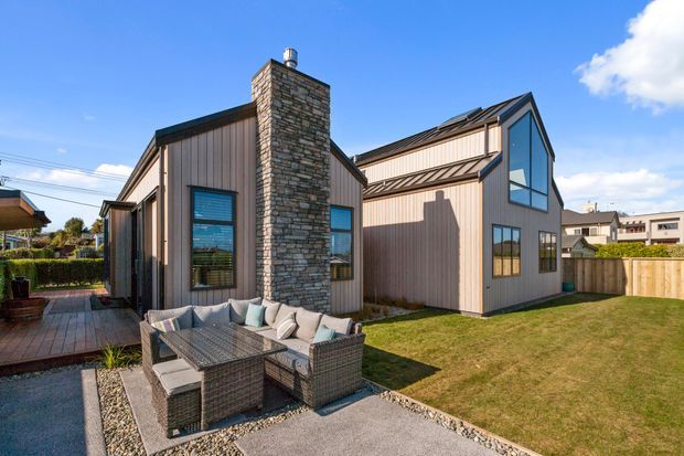 Exterior view of a contemporary multi-volume residential house with a steeply pitched roof, vertical metal cladding, a prominent natural-stone chimney, and large angular glazing. A paved patio with outdoor seating sits beside a lawn and timber boundary fence.