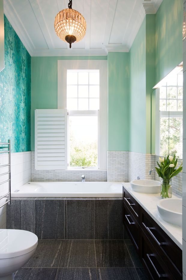 Bright, elegant bathroom with a freestanding-style built-in bathtub beneath tall multi-pane windows, a white vessel basin on dark timber cabinetry, white brick-look wall tiling, mint-green painted walls, patterned turquoise feature wallpaper, and a decorative woven pendant light. The room includes a towel warmer and toilet, with a refined traditional architectural character.
