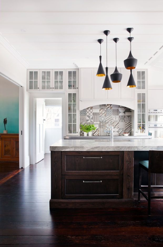Bright, refined kitchen interior featuring a large dark timber island with a pale stone benchtop, white paneled cabinetry with glazed display doors, and a decorative patchwork-pattern tiled splashback. A cluster of black sculptural pendant lights hangs above the island. Dark timber flooring and an adjoining darker-toned room are visible at the left.