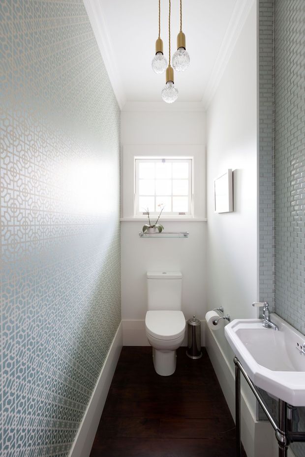 Narrow, bright bathroom with a toilet and traditional white pedestal-style basin, dark timber flooring, pale grey-white walls, patterned geometric wall tile on one side, small glazed window, and a textured pale blue-grey tile wall. Three pendant lights with brass-toned fittings and faceted glass shades hang from a white coffered ceiling.
