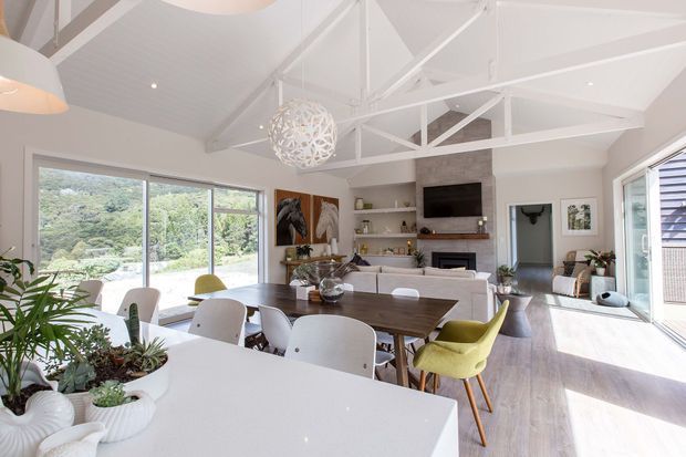 Bright open-plan living, dining and kitchen interior with a white island benchtop in the foreground, a large timber dining table, generous glazing, and a lounge area arranged around a fireplace and television. White-painted exposed roof trusses and a high vaulted ceiling create an airy pavilion-like space, with indoor plants and contemporary furnishings adding warmth.