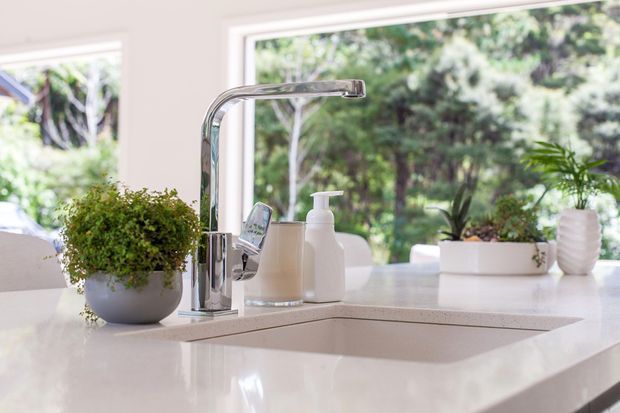 Bright contemporary kitchen sink area with a polished chrome gooseneck tap, integrated rectangular sink, white stone-look benchtop, and white soap dispensers. Large windows overlook lush greenery, while potted plants add a natural accent.