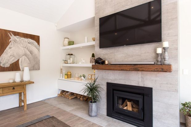 Bright contemporary living room featuring a wall-mounted flat-screen television above a black-framed fireplace with timber logs, set into a large light grey tiled feature wall. A floating white display cabinet and open shelves provide storage, while a timber console, neutral decor, indoor plants, and a monochrome horse artwork add warmth.