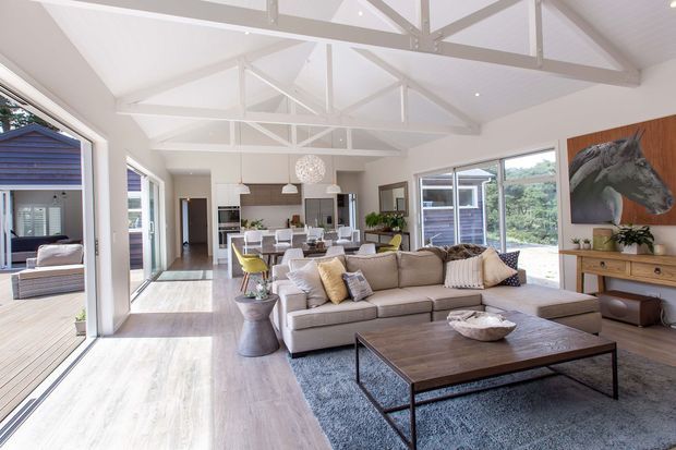 Bright, open-plan living room with a large sectional sofa, timber coffee table, media console, and dining and kitchen areas beyond. Extensive glazing opens toward outdoor spaces, while exposed white-painted roof trusses and a vaulted ceiling create an airy pavilion-like interior. Neutral furnishings, pale timber flooring, and a large horse artwork add warmth and character.