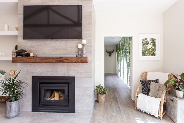 Bright contemporary living room featuring a wall-mounted television above a black fireplace set into a large grey stone or concrete-look feature wall. A timber mantel, light wood-look flooring, rattan armchair, neutral textiles, indoor plants, and artwork create a warm modern interior. A hallway with glazed or reflective doors is visible beyond the room.