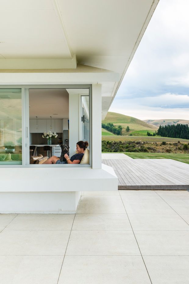 Exterior view of a minimalist contemporary home with broad white eaves, large aluminium-framed glazing, a pale tiled patio, and a timber deck overlooking rolling rural hills. A living area with a seated person is visible through the window.