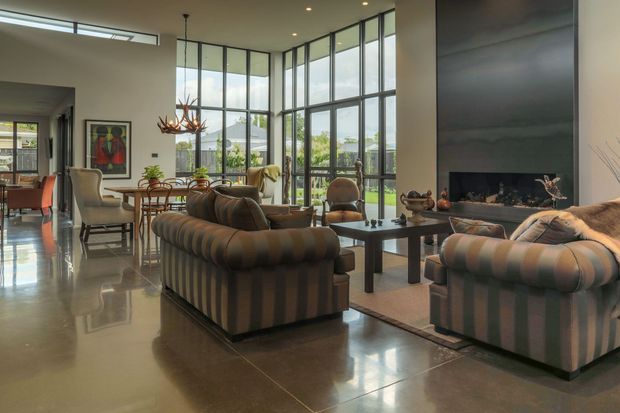 A spacious, refined living room with floor-to-ceiling black-framed glazing, polished concrete flooring, and a dramatic dark feature wall. Two large striped sofas face a low rectangular coffee table and fireplace/media wall, while a dining area and kitchen are visible beyond. A sculptural antler-style pendant light and curated furnishings add character to the open-plan interior.