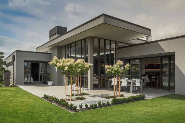Contemporary single-storey residence with a flat-roofed, geometric form, dark-framed floor-to-ceiling glazing, pale rendered walls, and a covered outdoor entertaining area. The landscaped foreground includes a manicured lawn, paved terrace, clipped garden edging, and young ornamental trees. Interior dining and kitchen areas are visible through the glazing.