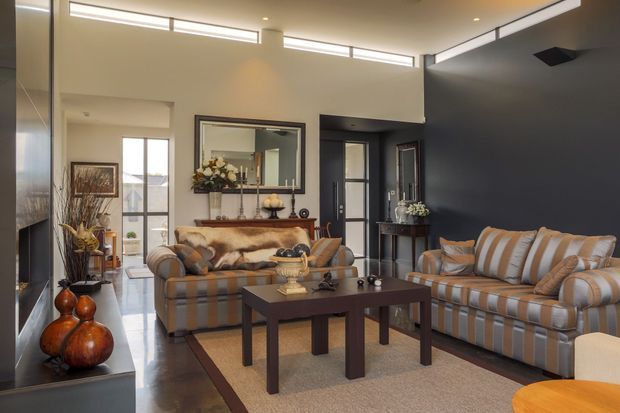 A refined living room with a high ceiling and clerestory windows, featuring striped upholstered sofas, a dark timber coffee table, a large framed mirror, decorative console styling, and a dark charcoal feature wall. The room has polished dark flooring, warm neutral furnishings, and abundant natural light.