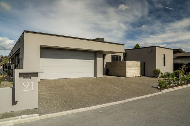 Street-facing view of a contemporary single-storey residence with a flat roof, minimalist pale rendered facade, large integrated double garage, covered entry, textured concrete driveway, and a low rendered boundary mailbox wall marked with the house number 21. A darker rendered side wing and screening wall define the entry courtyard.
