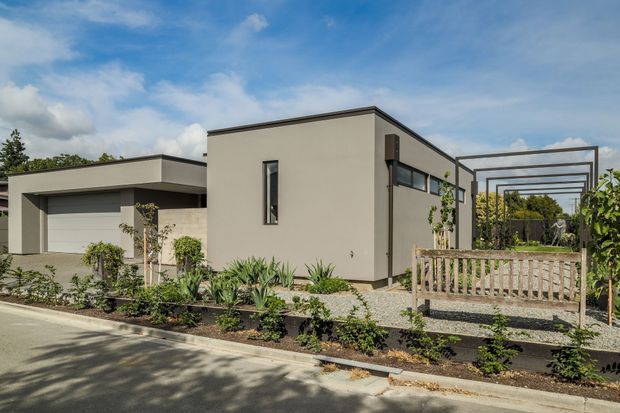 A contemporary single-storey residential home viewed from the street, with a minimalist rendered facade, flat rooflines, narrow vertical and horizontal windows, and an attached single-car garage. The landscaped frontage includes gravel, low planting, a timber pedestrian bridge or deck edge, and a dark metal pergola structure extending beside the house.