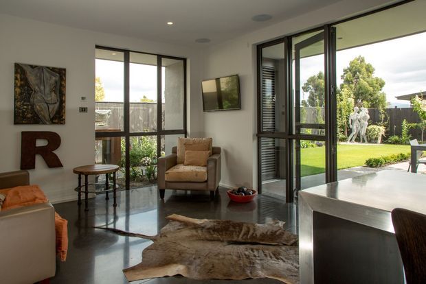 Bright contemporary living area with polished concrete flooring, floor-to-ceiling dark-framed glazing, and views to a landscaped garden. The room includes a tan upholstered armchair, sofa edge, side table, wall-mounted television, artwork, and a large sculptural cowhide rug. A sleek kitchen island or peninsula is visible in the foreground.