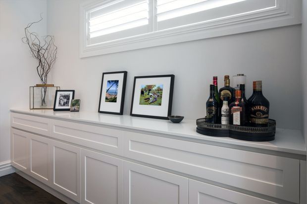 A bright, refined interior vignette featuring a long white built-in sideboard with recessed panel cabinetry, framed photographs, a decorative branch arrangement, and a tray of liquor bottles. The cabinetry sits beneath a wide white-framed louvred window, with grey walls and dark timber flooring.