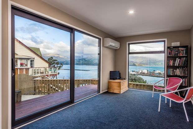 A bright waterfront-facing room with expansive floor-to-ceiling sliding glazing and a large picture window overlooking a bay, hills, and nearby homes. The room has dark blue carpet, warm taupe walls, a white ceiling with recessed lighting, a wall-mounted split air conditioner, a small timber TV cabinet, open bookshelves, and two vivid pink modern lounge chairs. A timber deck is visible beyond the sliding doors.
