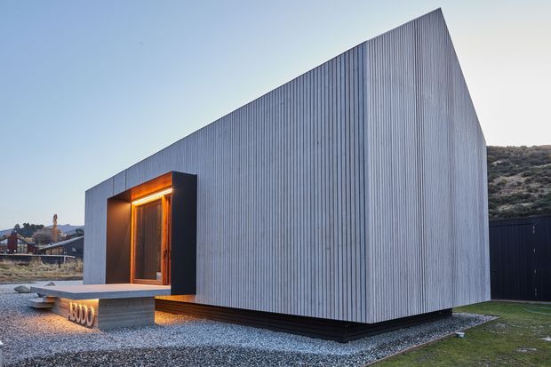 A compact contemporary single-storey building with a sharply folded gable-like roof form and vertically ribbed light-grey metal cladding. A dark recessed entry is illuminated with warm lighting, above a concrete step and gravel forecourt. The building is set in a sparse residential landscape with surrounding hills and neighboring structures.
