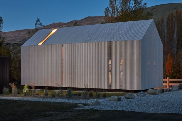 A compact contemporary gable-roofed building clad in vertically ribbed grey metal, set in a landscaped gravel and stone setting against a rugged hillside. Narrow vertical windows and a long roof skylight provide warm illumination at dusk.